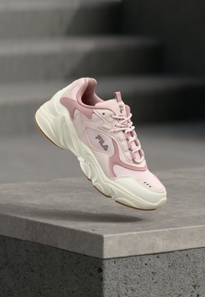 Baskets FILA rose clair et blanches flottant au-dessus d'un rebord en béton avec un arrière-plan gris flou.