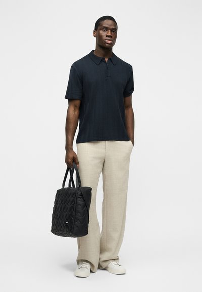 Άνδρας με μαύρο πικέ πόλο πουκάμισο, μπεζ παντελόνι με φαρδιές παντελόνι, λευκά αθλητικά παπούτσια, κρατώντας μια μαύρη τσάντα tote με ραφές, στέκεται μπροστά σε λευκό φόντο.