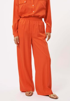Blouse orange avec deux poches poitrine, associée à un pantalon orange ample. Tissu lisse, taille élastique et sandales plates.