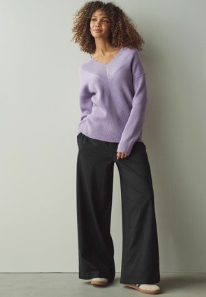 Lila gestrickter Pullover mit V-Ausschnitt und Rippstruktur, kombiniert mit schwarzen weit geschnittenen Hosen mit Nadelstreifenmuster und hellen Schuhen.