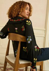 Különböző göndör hajú nő oldalra ül egy fából készült széken, fekete virágmintás hímzett blúzt és sötétkék nadrágot visel egy világos szobában.