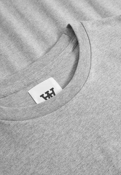 T-shirt en coton gris avec un col rond, des bords côtelés et une étiquette noire avec des lettres stylisées. Tissu doux et texturé.