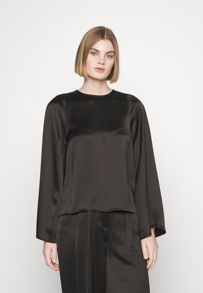 Femme avec des cheveux blonds lisses jusqu'aux épaules, portant un haut à manches longues en soie noire ample et un pantalon assorti, sur fond blanc.