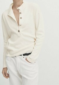 Persona che indossa una maglietta henley a maniche lunghe color crema con quattro bottoni e pantaloni bianchi con una cintura intrecciata nera, una mano in tasca.