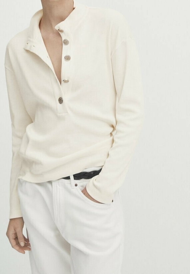 Persona che indossa una maglietta henley a maniche lunghe color crema con quattro bottoni e pantaloni bianchi con una cintura intrecciata nera, una mano in tasca.