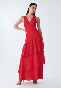 Red sleeveless dress with a V-neckline, featuring layered ruffles in an A-line shape, made from a smooth fabric, with a flowing skirt.