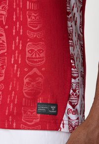 Rode shirt met textiel met een structuur, versierd met tribale maskermotieven en pijlen. Het heeft een zwart label met "Authentic Apparel" aan de zijkant.