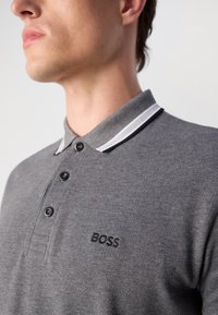 Grå polo skjorte med teksturert overflate, svarte knapper og kontrastfarget hvit stripet krage. Har et brodert "BOSS"-logo på brystet.