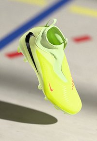 Cizme de fotbal galben strălucitor cu swooshes negre și roșii Nike, guler tip șosete și crampoane pe talpă, prezentate în aer, deasupra unei suprafețe gri.