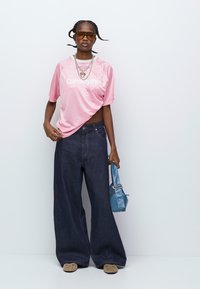 Lyserød oversized grafisk t-shirt, mørke bredbenede jeans, brune klipklapper og en blå håndtaske. Tilbehøret inkluderer solbriller og lagdelte halskæder.