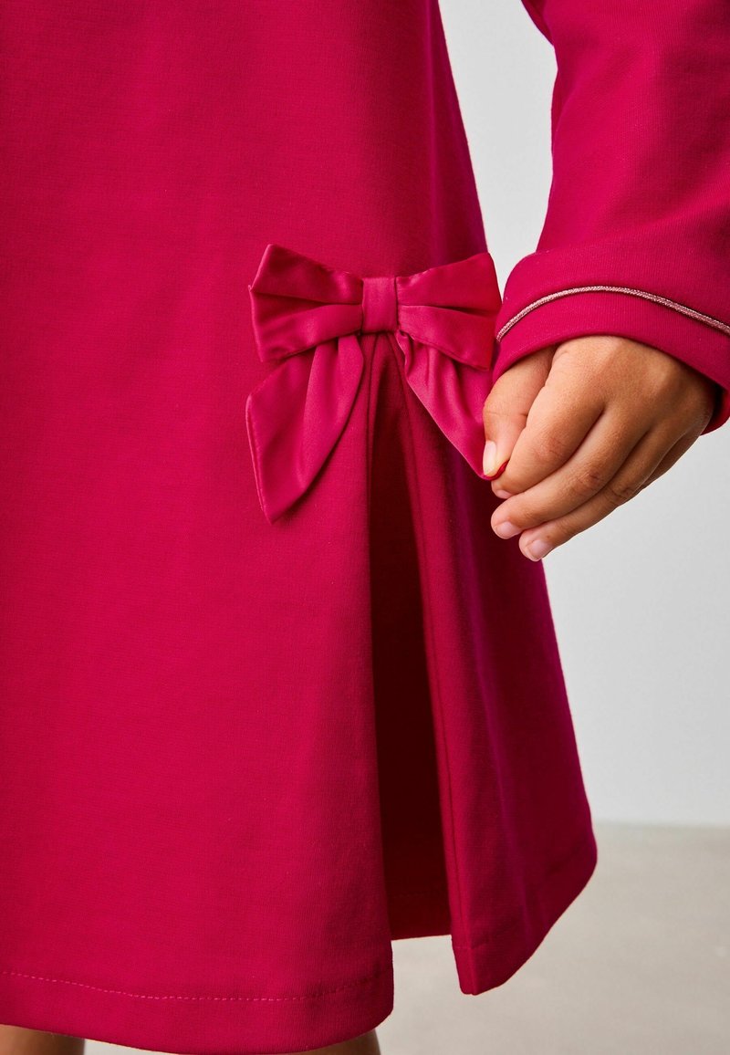 Main d’enfant à côté d’une robe rose foncé avec un détail de nœud en tissu et une manche longue avec un liseré rose clair au poignet.