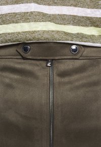 Olivgrüne Wildlederhosen mit einem Reißverschluss vorne, Druckknopfverschluss und einer glatten Textur. Darüber ist ein gestreifter, hellgrüner Pullover getragen.