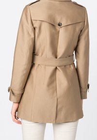 Manteau beige avec une taille ceinturée, des épaulettes et des détails de boutons. Fabriqué en tissu lisse, avec une coupe ajust�ée et une fente au dos.
