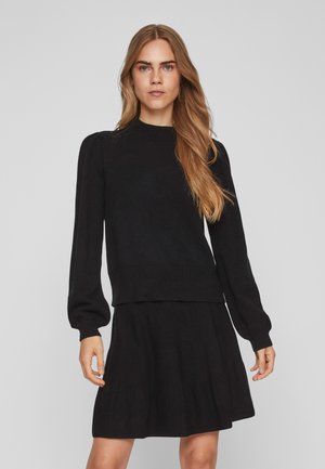 Pull noir à manches longues avec un col rond côtelé et des épaules légèrement bouffantes, porté avec une jupe assortie à pans superposés. Texture douce.