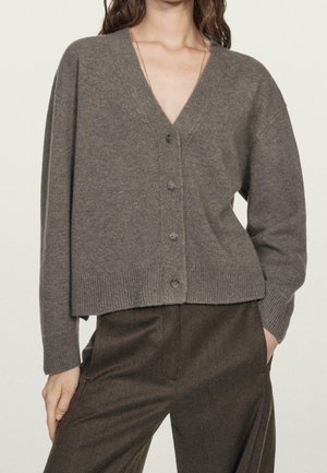 Vrouw die een bruine gebreide cardigan met V-hals en knopen draagt, samen met bijpassende bruine broek, met één hand in haar zak, tegen een effen achtergrond.