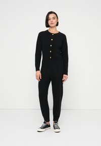 Μαύρη ολόσωμη φόρμα πλεκτή με ριμπ υφή, μακριά μανίκια και στρογγυλή λαιμόκοψη, με κουμπιά σε χρυσό τόνο κατά μήκος της μπροστινής πλευράς και ρεβέρ στα μανσέτα των ποδιών. Συνδυάζεται με αθλητικά παπούτσια.