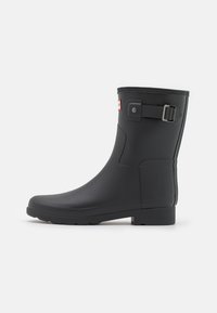 Bota de lluvia de goma negra con un caño texturizado, punta cuadrada, suela plana y un detalle de hebilla ajustable en el lateral. Acabado liso.