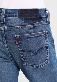 Primer plano de una persona que lleva vaqueros azules de mezclilla con un bolsillo trasero y una etiqueta roja visible de Levi's en el lado derecho.