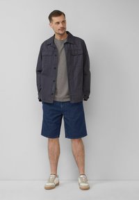 Donkerblauwe canvas jas met knoopsluiting, gecombineerd met een grijs t-shirt, denim shorts en crèmekleurige sneakers met bruine accenten.