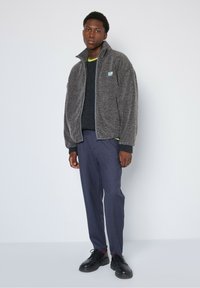 Graue Fleecejacke mit hohem Kragen, Reißverschluss und kontrastierendem grünen Akzent. Kombiniert mit marineblauen Hosen und schwarzen Schuhen.