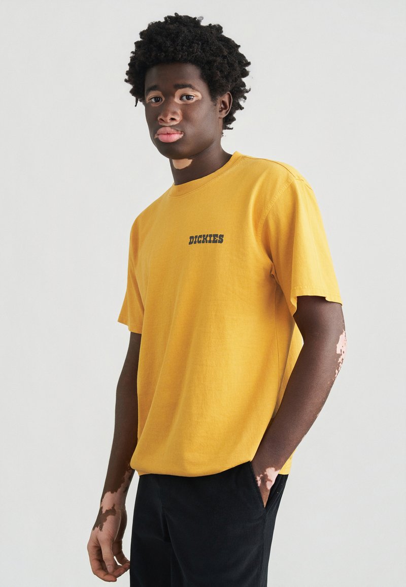 Κοντομάνικο, κίτρινο βαμβακερό μπλουζάκι με στρογγυλή λαιμόκοψη και μαύρο λογότυπο «Dickies» στο στήθος· συνδυασμένο με μαύρο παντελόνι.