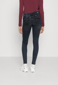 Calças skinny em denim de lavagem escura, com dois bolsos traseiros com abas angulares, combinadas com uma blusa de mangas longas cor bordô e sapatilhas brancas.
