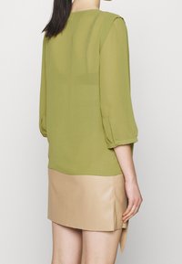 Olivgrübluse aus halbtransparentem Material mit Dreiviertelärmeln, geknöpften Manschetten und geradem Saum, kombiniert mit einem beige Lederrock.