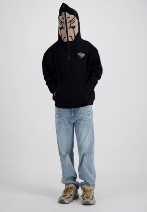 Person trägt einen schwarzen Kapuzenpullover mit Halb-Reißverschluss-Gesichtsmaske, die ein abstraktes beiges Gesicht zeigt, hellblaue zerrissene Jeans und beige Sneakers.