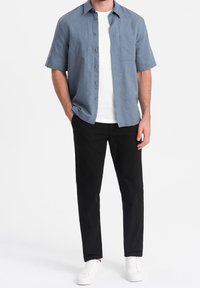 Man som bär en blå kortärmad skjorta över en vit t-shirt, svarta byxor och vita sneakers, stående med ena handen i fickan.