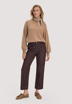 Blonde vrouw met een beige sjaaltrui, donkerbruine hoge taillebroek en bruine suède instapschoenen, staand voor een lichte achtergrond.