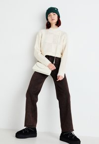 Suéter de malha creme com um padrão de rede texturizada, calças castanhas largas, sapatos plataforma pretos com detalhes brancos, e um gorro verde.