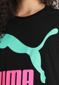 Camiseta negra con un gran logo de Puma en turquesa y el texto "PUMA" en rosa neón. Hecha de un tejido suave con una textura lisa.