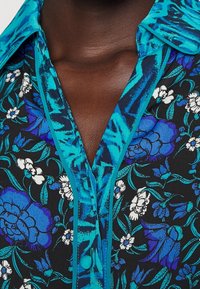 Camicia floreale con una base nera, decorata da motivi blu e bianchi, colletto turchese e chiusura a bottoni. Tessuto morbido con colori vivaci.