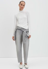 Weißes langärmliges Turtleneck, graue Hose mit geknotetem Stoff an der Taille und weiße Slipper vor einem hellen Hintergrund.