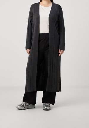 Persoon die een lange donkergrijze open cardigan draagt, een wit top, zwarte wijde pijpen broek en grijze sportschoenen, staande op een grijze vloer.