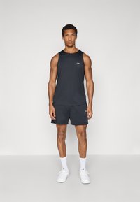 Schwarzes ärmelloses Sport-Tanktop und schwarze Shorts mit einem Logo auf der Brust. Getragen mit weißen Schuhen und Socken. Glatter Stoff, figurbetontes Design.