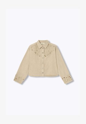 Chemise beige à manches longues avec col et boutons, ornée de broderies florales ajourées sur la poitrine et les poignets, posée à plat sur un fond blanc.