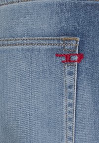 La tela de mezclilla azul claro presenta un primer plano de un bolsillo trasero con costuras visibles y un detalle de logotipo bordado en rojo.