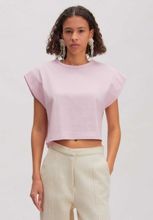 Vrouw met krullend haar die een lichtroze mouwloos topje en beige broek met hoge taille draagt, met grote hangende pareloorbellen.