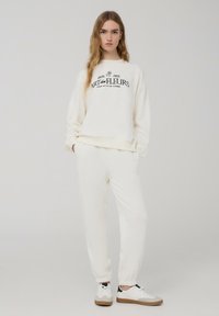 Felpa color crema e pantaloni jogger coordinati, con una stampa grafica nera. Tessuto morbido con polsini elastici e vestibilità rilassata. Sneakers bianche con dettagli beige.