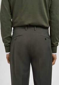 Back view of person wearing dark gray tailored trousers and a tucked-in olive green long-sleeve sweater with buttoned back pockets.