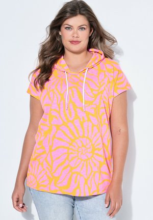 Femme portant un sweat à capuche à manches courtes rose et orange à motifs avec des cordons blancs, associée à un jean bleu clair, debout sur un fond blanc.