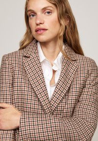 Blazer em padrão houndstooth castanho e branco, com lapelas à espanhola, tecido texturado, fecho de um só botão e detalhes nos punhos.