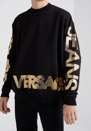 Personne portant un sweat-shirt noir avec un grand texte métallique doré "VERSACE" sur la poitrine et les manches, debout devant un fond uni.