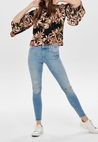Blusa nera floreale con maniche a campana, abbinata a jeans skinny strappati blu chiaro e sneakers bianche. Tessuto testurizzato con motivi a foglia.