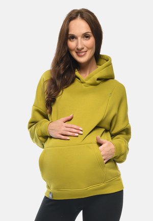 Donna sorridente in felpa verde con cappuccio, con la mano sul pancione, che dimostra un abbigliamento premaman con zip laterale per l'accesso all'allattamento.