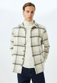 Knoopshirt in beige en zwart plaid stof, met een kraag, lange mouwen en een relaxte pasvorm, gedragen over een crèmekleurige trui met hoge hals.