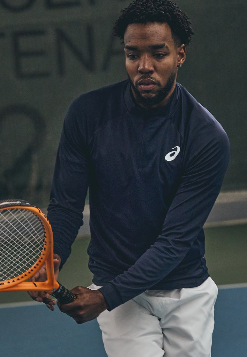 Navyblaues, langärmliges Sportshirt mit einem Viertelreißverschluss-Design und Logo auf der Brust, kombiniert mit weißen Shorts und einem orangefarbenen Tennisschläger.