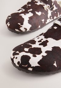 Zoccoli marroni e bianchi con motivo a fantasia, realizzati in morbido materiale di feltro, con forma arrotondata e superficie strutturata. Suola in gomma nera con leggera aderenza.