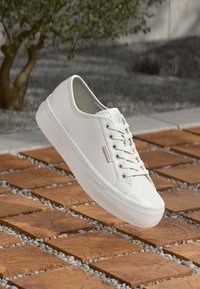 Weiße Calvin Klein Sneaker mit Schnürsenkeln, die über einem Holzboden mit Fliesen schweben, im Hintergrund sind ein Baum und Kies zu sehen.